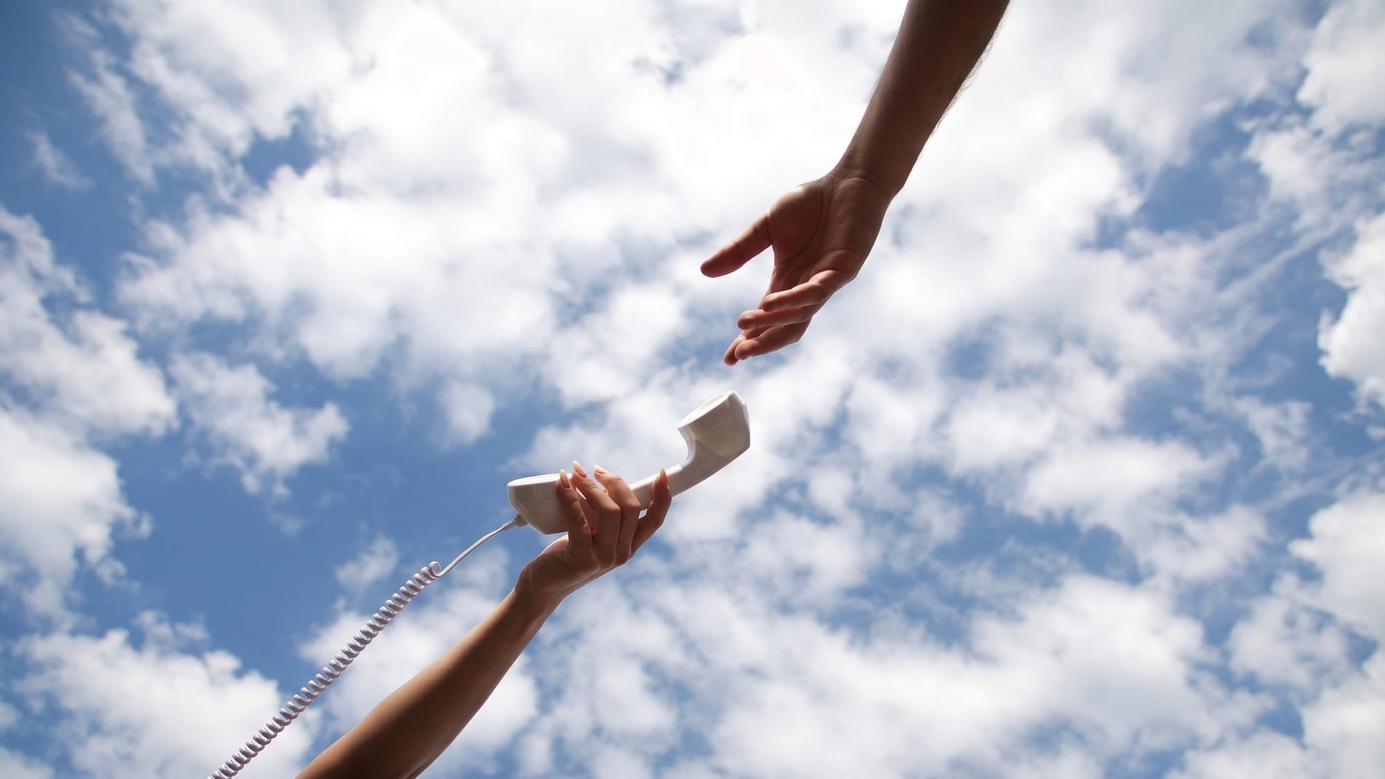A hand holds a white corded telephone receiver while a second hand reaches towards it from above, with a blue sky and clouds in the background.