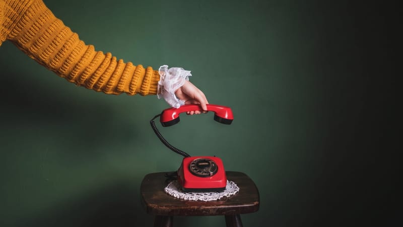 A hand in a mustard jumper with a lace cuff lifts the receiver of a red telephone from a wooden stool in front of a green wall.