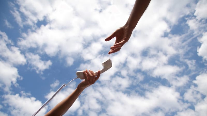 A hand holds a white corded telephone receiver while a second hand reaches towards it from above, with a blue sky and clouds in the background.